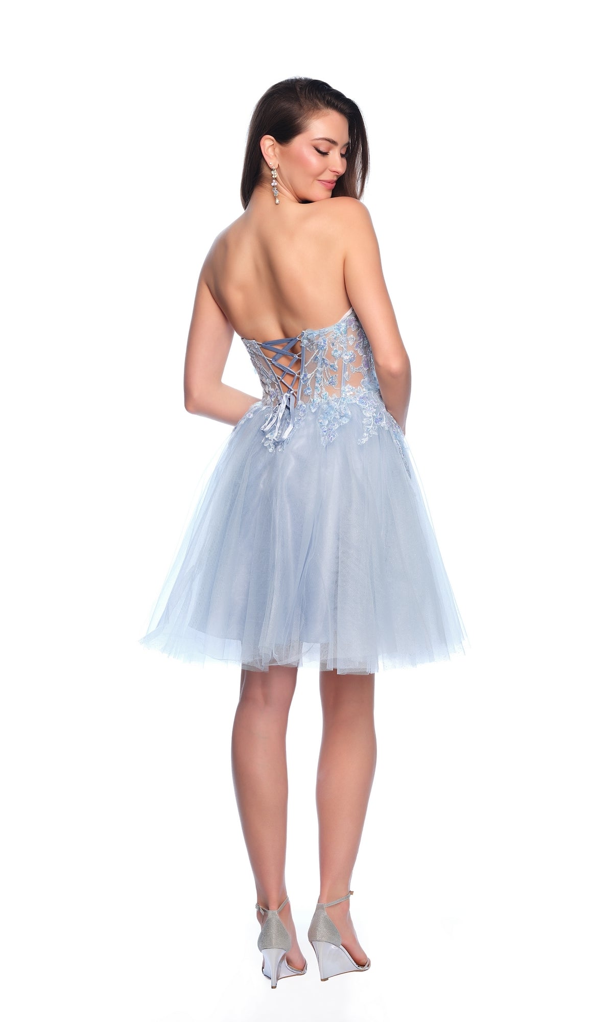 A woman stands with her back to the camera in the Sheer Glitter Homecoming Dress 11973, featuring a light blue strapless design, lace-up back, and tulle skirt.